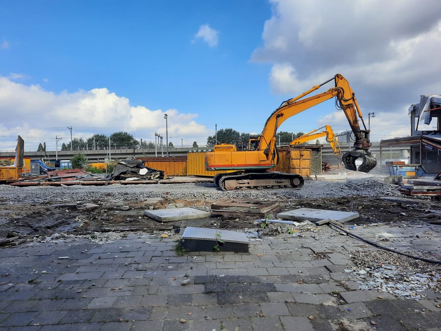 The image depicts a construction site with a yellow excavator positioned on gravel, actively lifting debris with its hydraulic arm. The debris includes broken pavement slabs, metal sheets, and various