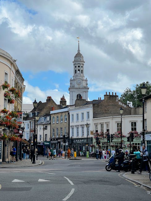 A busy street scene in a historic town, featuring a row of traditional Victorian-style buildings with shopfronts, some decorated with hanging flower baskets. Pedestrians walk along the pavement, and a