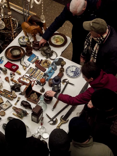 Inside a spacious warehouse or market hall filled with a large crowd of people browsing and shopping at various tables and stalls. The tables are covered with a wide array of items, including books, a